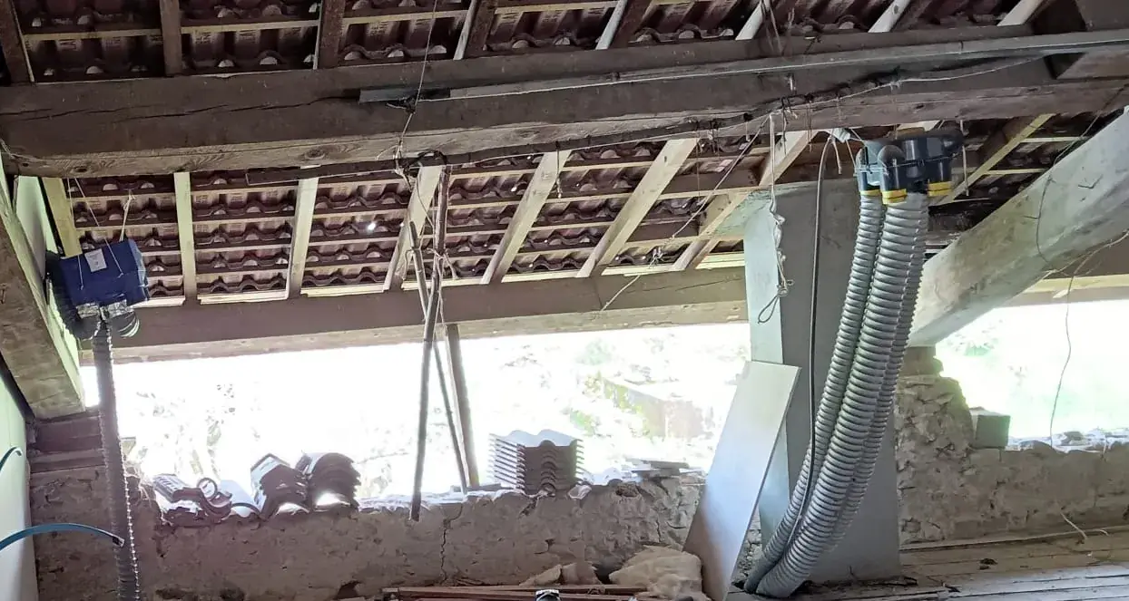 Toiture vue de l’intérieur : tuiles sur liteaux et chevrons, jour entre la toiture et un mur en pierre où des tuiles sont empilées