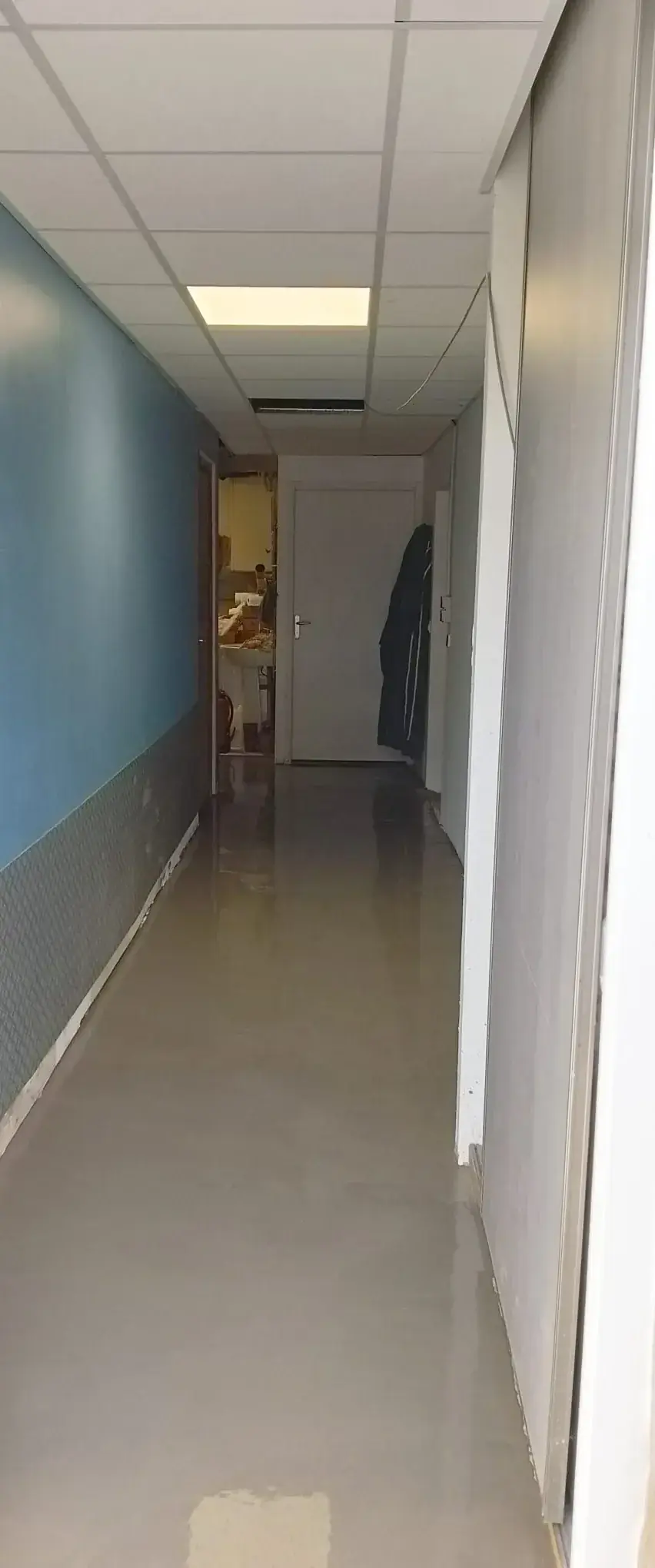 Couloir au sol gris lisse et brillant, mur bleu au-dessus d'un soubassement texturé, faux plafond à dalles
