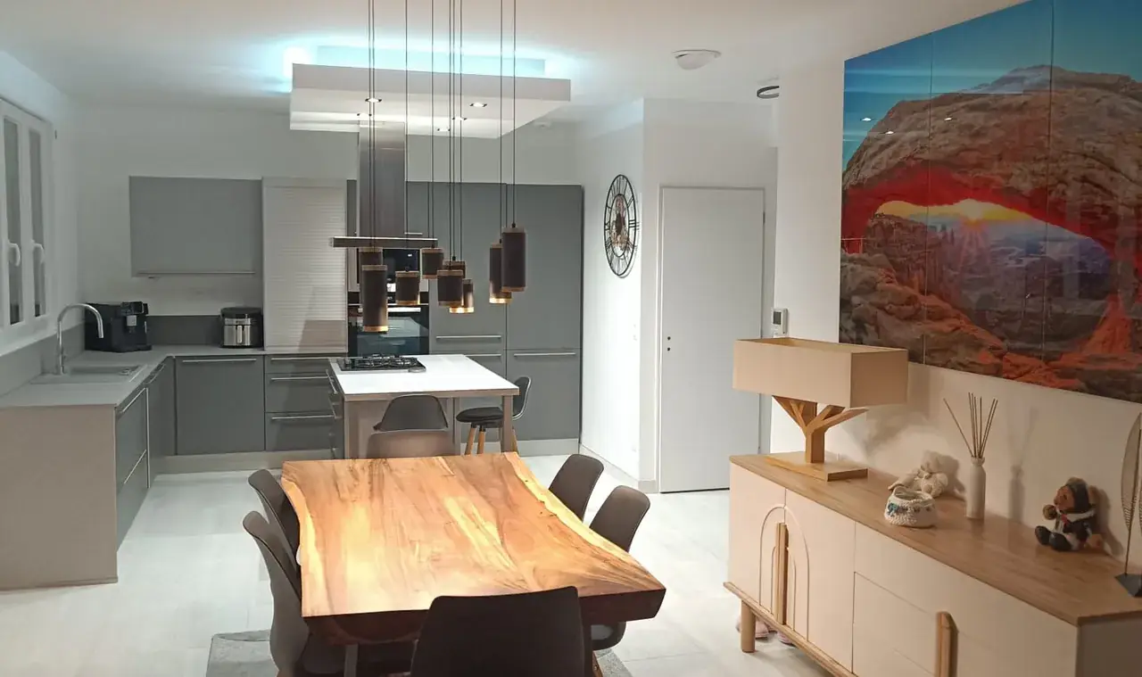 Cuisine ouverte avec îlot sous un caisson lumineux au plafond ; table en bois au premier plan, buffet clair à droite