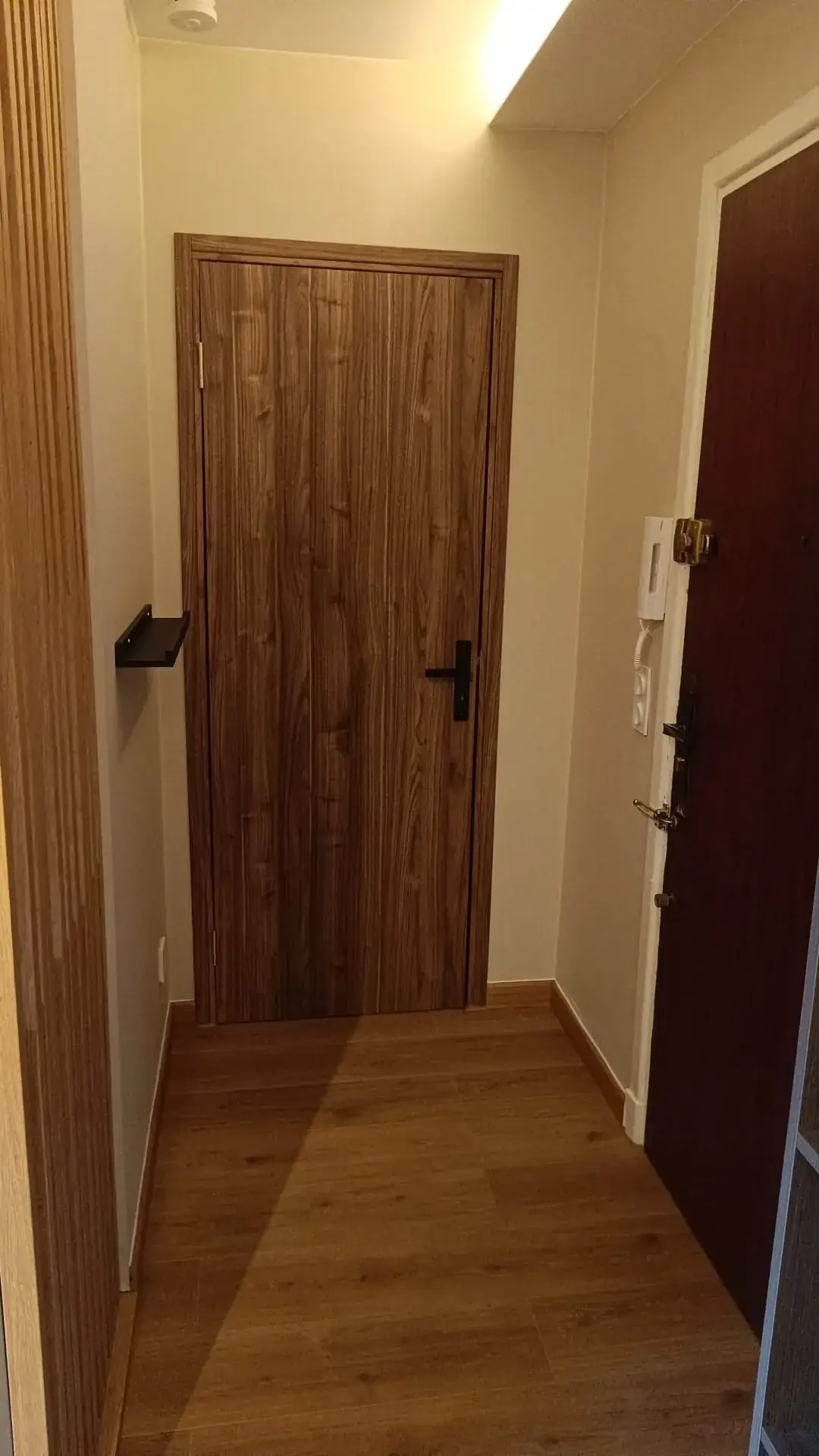 Entrée : porte à décor bois au fond, sol en lames de teinte bois, porte d'entrée rouge sombre sur la droite