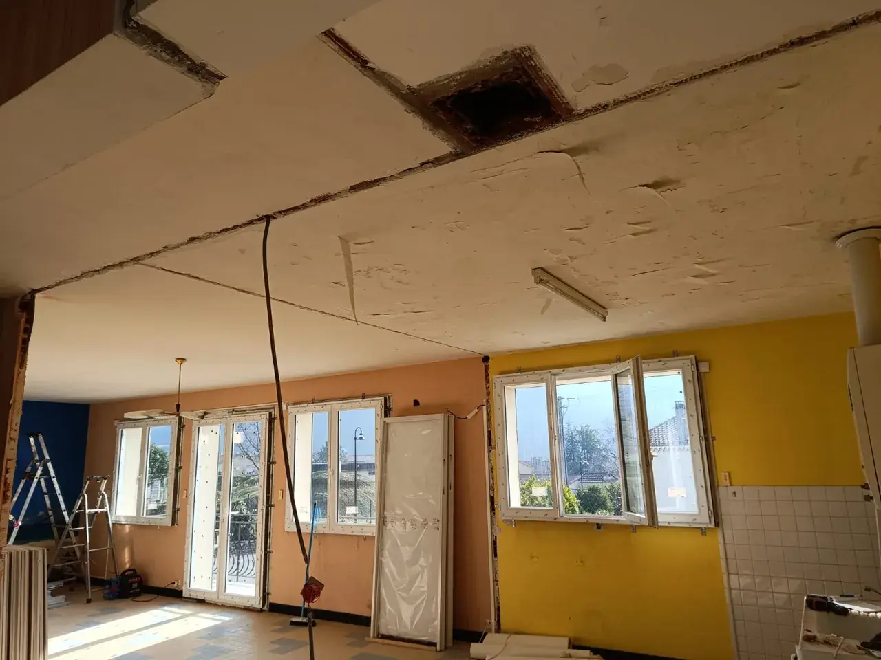 Plafond qui s'écaille par lambeaux, joints ouverts et ouverture carrée ; fenêtres et porte-fenêtre blanches dans les murs saumon et jaune
