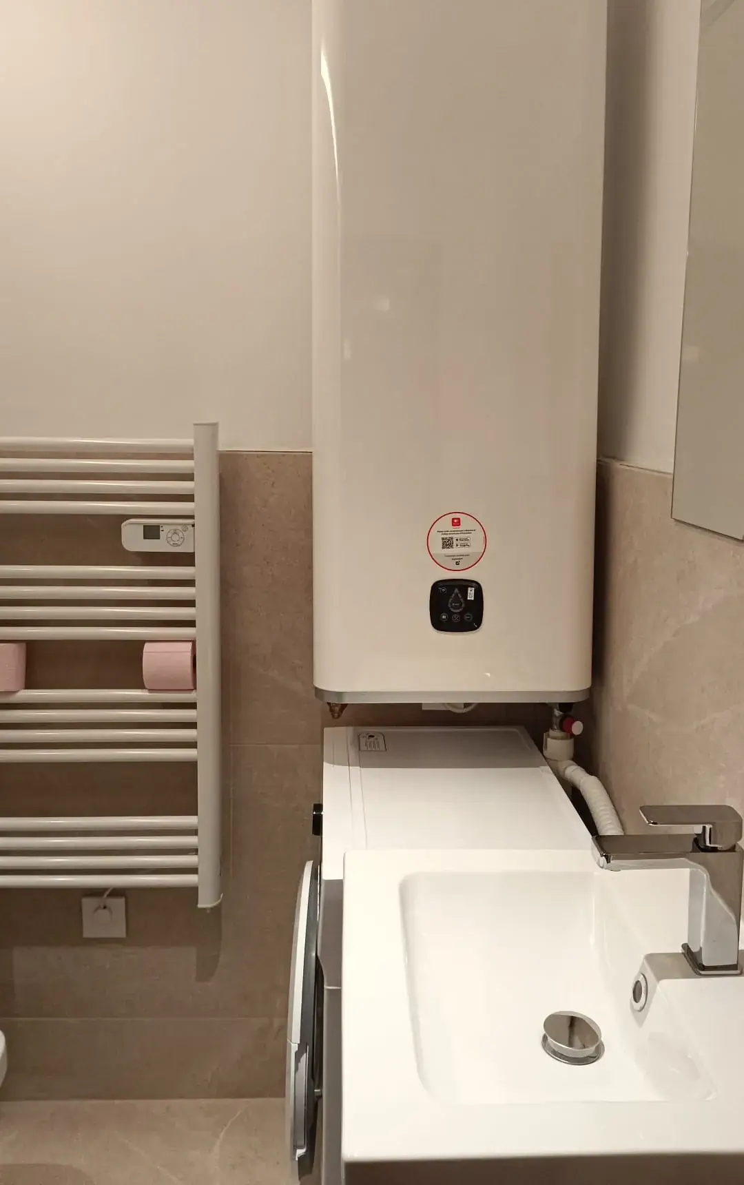 Chauffe-eau mural au-dessus d'un lave-linge, sèche-serviettes à gauche et vasque au premier plan, carrelage beige en partie basse des murs