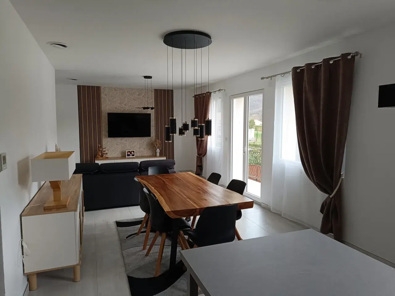 Séjour : table en bois et suspension à cylindres noirs ; télévision sur un pan de mur à motif de feuilles, entre deux panneaux à lames verticales couleur bois