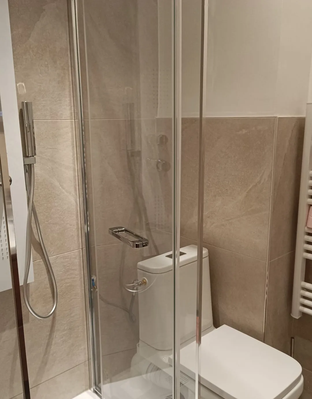 Douche fermée par une paroi vitrée coulissante, WC et sèche-serviettes, murs en carrelage beige