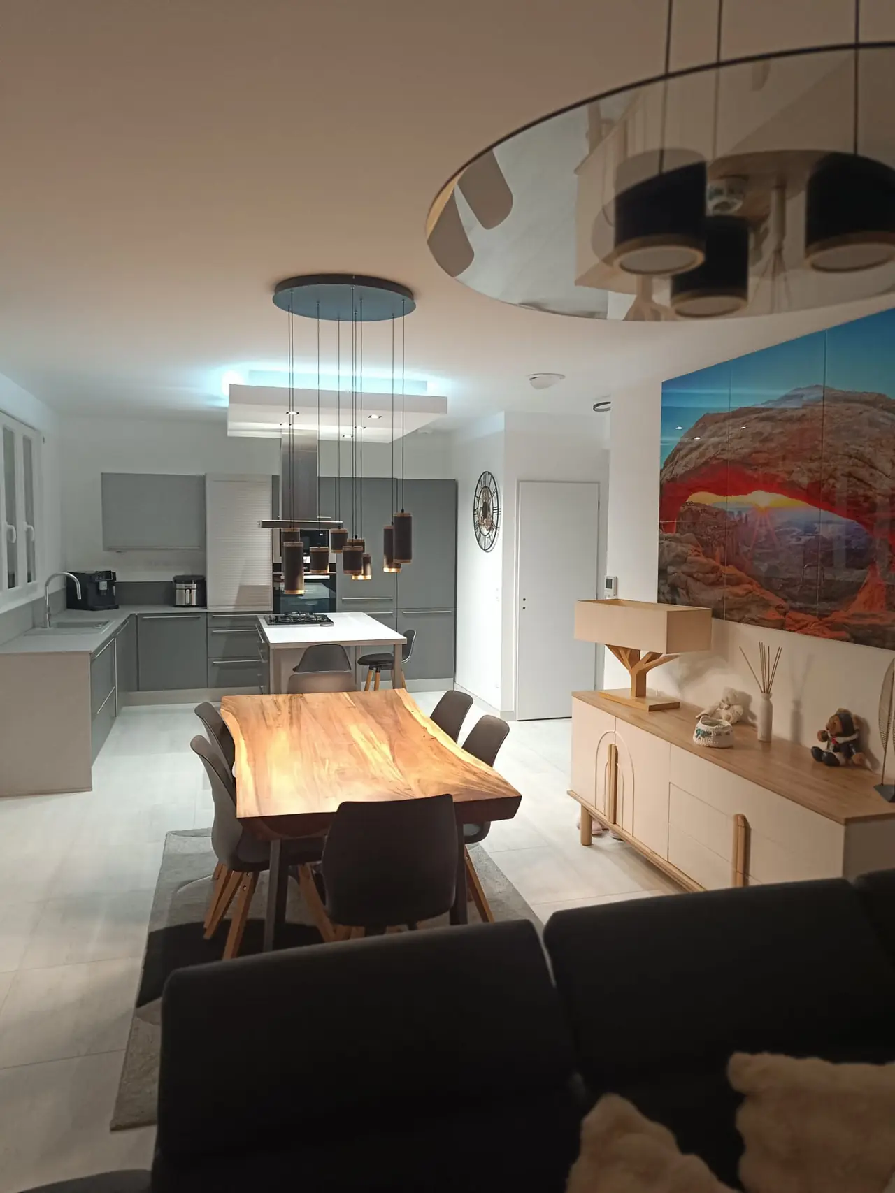Pièce de vie terminée : cuisine ouverte avec îlot sous un caisson lumineux, table en bois massif sous une suspension, buffet clair au pied d'un grand tableau